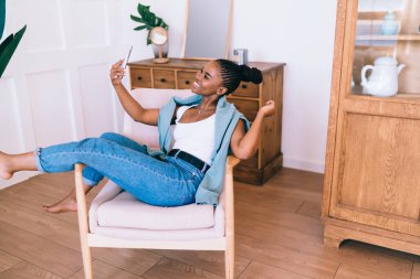 Pozitif genç Afro-Amerikalı kadın, gündelik giysiler içinde koltukta oturuyor ve evde akıllı telefondan selfie çekerken gülümsüyor.