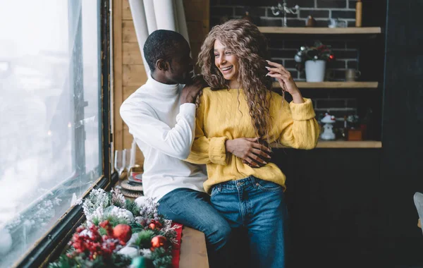 Happy couple in cozy interior at winter.Black man affectionately adjusts womans curly hair. She smiles brightly, clad in a yellow knit sweater, feeling the holiday spirit amidst festive decorations