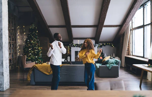 Joyful dance of Black man and woman in festive loft with Christmas tree. He shares dance step with her, enjoying the rhythm in a bright yellow blouse and blue jeans, embodying the holiday spirit