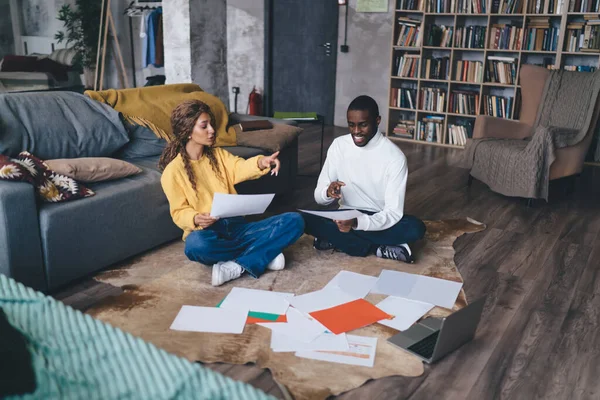 Young African-American couple engaged in planning, sitting on the floor amidst papers, laptop nearby, sharing ideas and smiling during a productive home meeting.