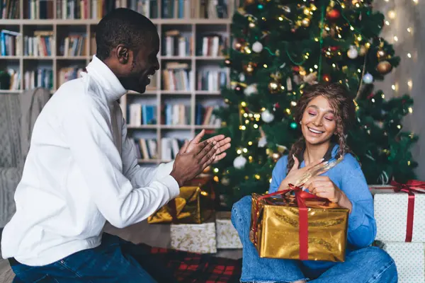 An elated Caucasian woman unwrapping a golden gift as an African American man in a white turtleneck joyfully claps, in a festive room with a Christmas tree and a wall of bookshelves