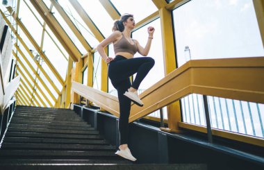 20 'li yaşlarda beyaz kadın merdiven tırmanışı yapıyor, spor bir üst ve siyah tayt giyiyor, şehir merdiven boşluğunda