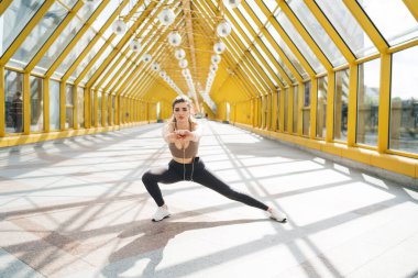 20 'li yaşlarda, saydam cam duvarları ve tavanı olan geniş sarı bir yaya köprüsünde yan atlama pratiği yapan beyaz bir kadın. Kahverengi spor bluzlu, siyah taytlı, beyaz spor ayakkabılı.