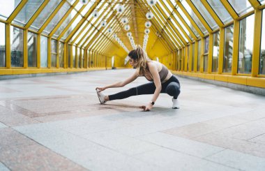 20 'li yaşlarda genç beyaz bir kadın parlak, sarı bir yaya köprüsünde esneme egzersizi yapıyor. Kahverengi spor sutyen, siyah tayt ve beyaz spor ayakkabı giyiyor. Müzik dinliyor.