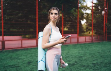 Yoga minderi ve su şişesiyle zinde genç bir kadın parkta akıllı telefon, teknoloji ve fitness konsepti kullanıyor.