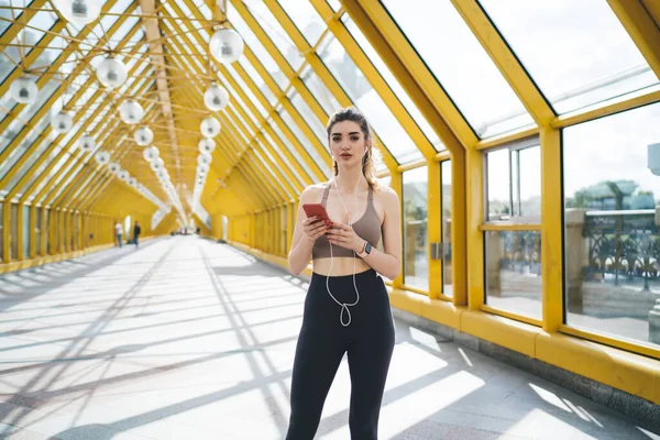 Tech-savvy young Caucasian woman in workout attire using her smartphone in a sunny yellow pedestrian overpass, with earphones and a fitness band