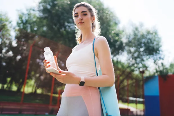 stock image Sporty woman with yoga mat holds water bottle and smartphone, ready for fitness session in park