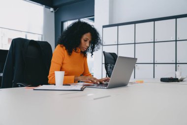 Canlı turuncu kazaklı odaklı girişimci özenle dizüstü bilgisayarla çalışıyor, modern ofis ortamında iş stratejileri planlıyor.