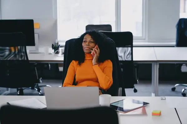 Focused Black businesswoman in orange turtleneck working on laptop in modern office setting, displaying professionalism and dedication with a pleasant demeanor.