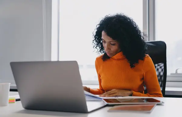 Focused African American administrative professional in her 30s coordinating remote office operations, deeply engrossed in paperwork, with a laptop and tablet on her desk in a well-lit co-working