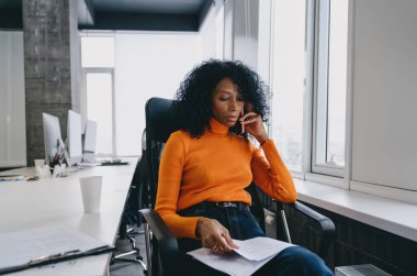 Parlak turuncu kazaklı endişeli bir çevre analisti, modern bir şirket ofisinde sürdürülebilir uygulamalar hakkında önemli bir telefon brifingi sırasında araştırma raporlarını inceliyor..