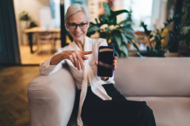 Kanepede oturan ve ofiste çalışırken akıllı telefon ekranını işaret eden mutlu son sınıf bayan girişimcinin yumuşak odağı.