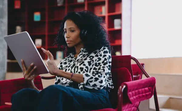 Focused Black businesswoman in her 30s, wearing a black and white animal print blouse and headphones, working intently on her tablet in a vibrant office lounge with red chairs and bookshelves.
