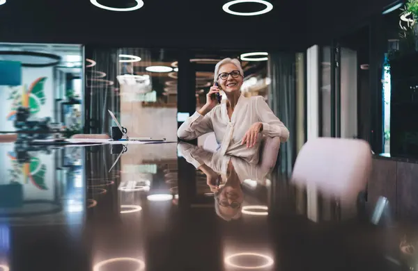 Low angle of positive businesswoman in classy outfit smiling while having phone conversation communicating on cellphone during business work in modern workspace