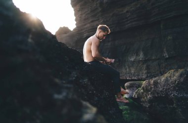 Çıplak ayaklı, pantolonlu, kayalık uçurumun yamacında oturan ve güneşli bir günde tatilin tadını çıkaran genç bir erkeğin yan görüntüsü.