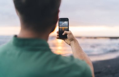 Arka planda, gece bulanık arka planda dikilirken deniz manzaralı günbatımının fotoğrafını çeken sıradan giysili, sıradan bir insanın görüntüsü var.