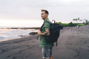 Sırt çantasıyla gezen genç bir erkek gezginin ıslak kumlu sahilde durup bulanık arka planda dalgalanan okyanusa bakışları.