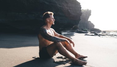 Çıplak ayaklı, şort ve kollu, kumlu sahilde güneşli bir günde kayalık arazide ve dağ yamacında oturan çıplak ayaklı genç bir erkek.