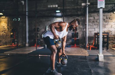 Tıraşlı rastalı ve spor kıyafetli genç Afrikalı sakallı erkek sporcunun spor salonunda antrenman yaparken iki elinde ağır kettlebell kaldırışının vücut tarafı.