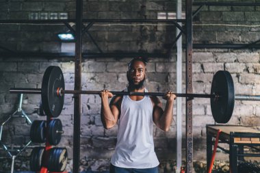 Sporcu, spor kıyafeti giymiş, spor salonunda ağır halterlerle ağırlık kaldıran güçlü bir Afrikalı Amerikalı.