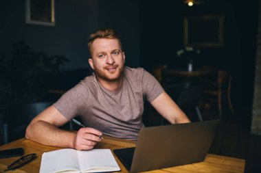 Mutlu sakallı erkek öğrenci, evden uzakta taşınabilir bilgisayarda ödevlerini yaparken defter ve kalemle gülümsüyor.