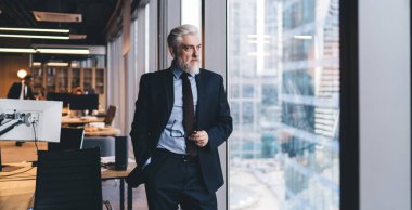 Konsantre yaştaki erkek yönetici resmi kıyafetler giyer. Pencere kenarında el ele durur ve modern çalışma alanına bakar.