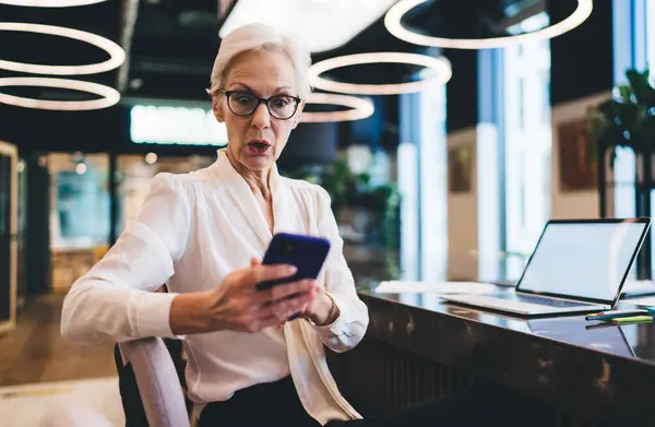 Elderly businesswoman looking at screen of mobile phone in disbelief and text message received in free time in office while sitting at table with laptop