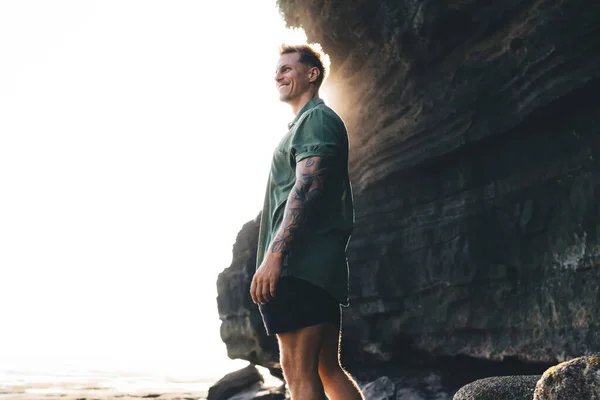 Low angle side view of young male traveler in casual clothes standing on rocky cliff and looking away with smile