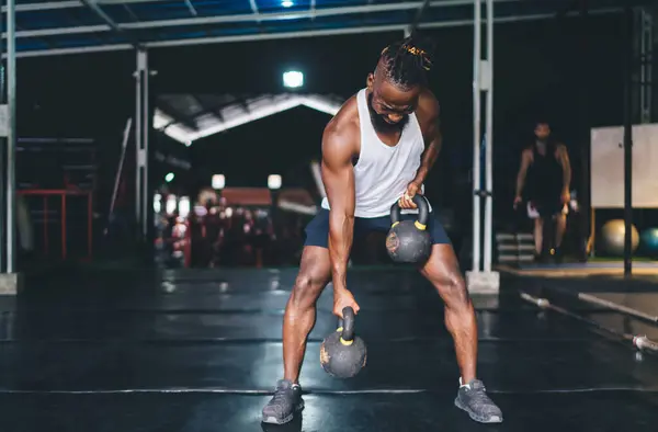 Full body of strong African American bearded male athlete in shaved dreadlock and sportswear lifting heavy kettlebells with both hands while working out in gym on dark background