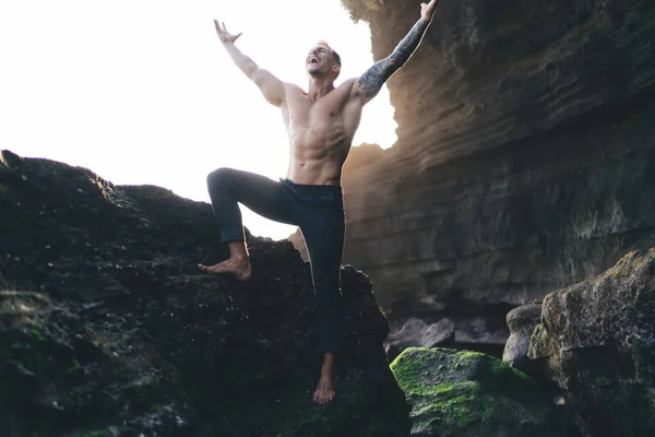 Full body of young shirtless male in casual pant standing on rocky cliff with opened arms and opened mouth while shouting and celebrating victory in daylight