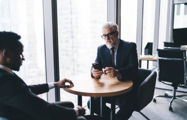 Resmi kıyafet giyen, akıllı telefon kullanan ve ahşap masada oturan, modern çalışma alanının penceresinin yanında çalışan ciddi bir kıdemli adam.