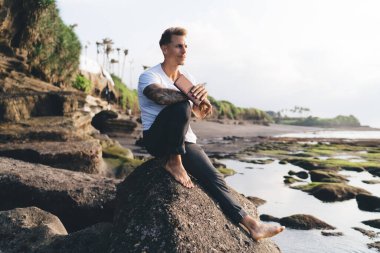 Nehrin kayalık kıyılarında güneşli bir günde kameraya bakarken, günlük giysiler içinde kitap tutan sakallı bir erkek.