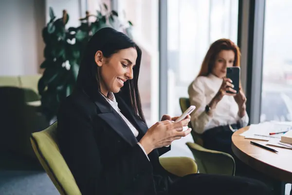 Side view of smiling female entrepreneur in formal clothes sitting at table with female colleague and browsing mobile phone while working in modern office