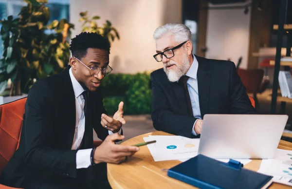 Busy business partners in classy outfits with glasses on working with documents and netbook in workplace while black man showing message on smartphone