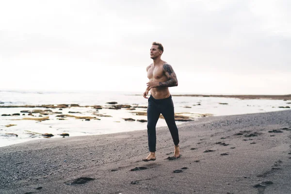 Full body of shirtless young male athlete running along sandy beach and enjoying morning fitness workout alone on seashore against blurred background