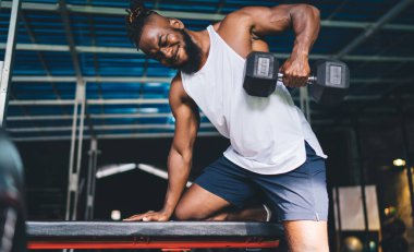Spor kıyafetli Afrikalı sakallı güçlü erkek sporcunun düşük açısı spor salonunda egzersiz yaparken tek diziyle ve koluyla ağırlık kaldırırken.