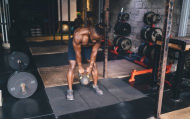 Güçlü Afrika kökenli Amerikalı sporcu spor salonunda spor yaparken ağırlık kaldırıyor ve öne eğiliyor. Spor aletleri duvarın yanında.