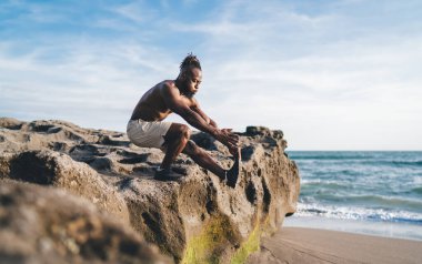 Güneşli bir günde bulutlu mavi gökyüzüne karşı dalgalanan denizin yakınındaki kayalık sahilde antrenman yaparken esnek bacaklı ve elleri antrenmanlı kalçaların üzerinde oturan üstsüz bir erkek.