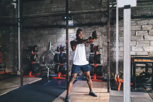 Full body of strong African American bearded male in sportswear standing straight and swinging heavy kettlebell with both hands during workout in gym with block wall