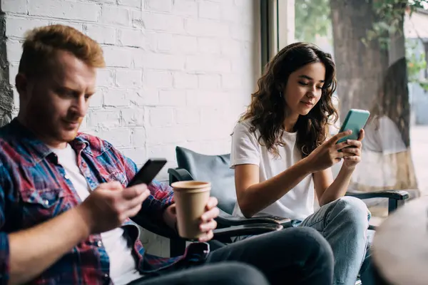 stock image Young man with hot drink in paper cup sitting with woman in cafeteria near window and using smartphones messaging during coffee break