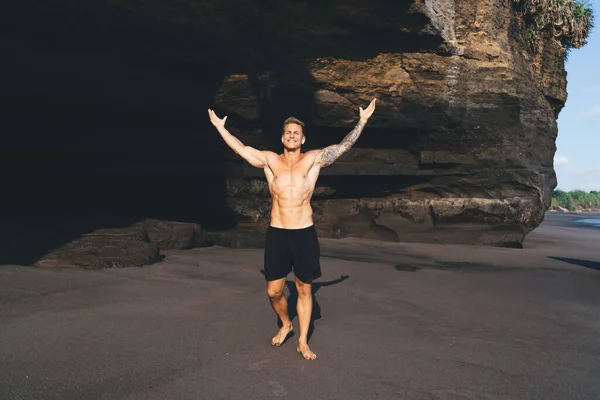 Full body of young shirtless muscular male with raised arms and closed eyes standing on sandy beach by rock celebrating joyful victory while enjoying nature in daylight