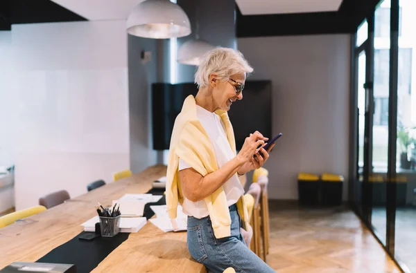 Positive senior female in casual outfit and eyeglasses with smartphone smiling and looking at camera while leaning on wooden table in modern office