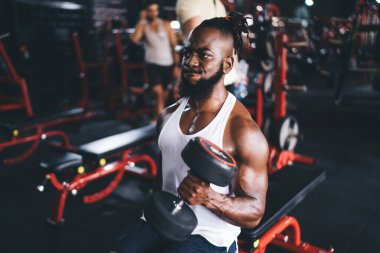 Kas yığını Afrikalı Amerikalı sakallı genç erkek sporcu güçlü bir yüz ifadesi takınarak halter kıvrımlı ağırlık kaldırıyor ve modern spor salonundaki bankta bulanık arka planda oturuyor.
