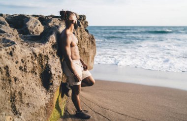 Gömleksiz, sakalsız, şortlu, siyah bir erkek. Bir bacağı havada, kumlu sahildeki kayalık tepede dinleniyor. El sallıyor ve tatilin tadını çıkarırken başka yere bakıyor.