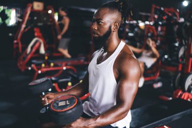 Yüksek açılı genç sakallı Afrikalı Amerikalı erkek sporcunun spor kıyafetleri içinde halterleri dizlerinin üzerinde bankta otururken bulanık arka plana bakarken görüntüsü.