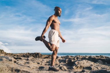 Mavi gökyüzüne karşı kayalık deniz kıyısında ısınırken kuadriseps germe egzersizi yapan kendine güvenen Afrikalı Amerikalı erkek.