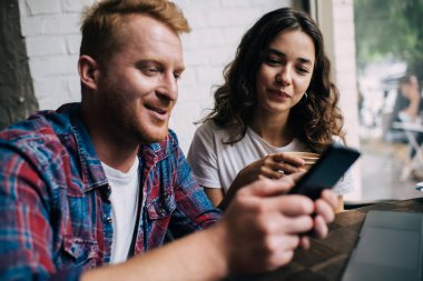 Modern kahve dükkanında masada otururken sıcak çay içen gülümseyen kadına akıllı telefondan bilgi veren genç adam.