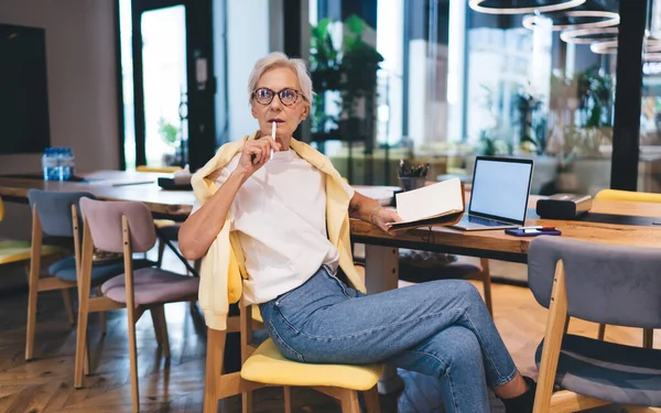 Pensive elderly female entrepreneur in casual clothes and glasses sitting at table with laptop while thinking about project and taking notes in notebook