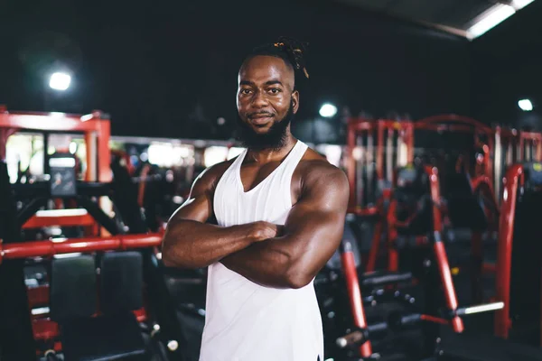 Portrait of strong African American sportsman in sportswear smiling and looking at camera while standing with folded arms in modern gym with sport equipment