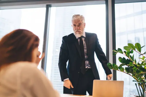 Low angle of annoyed male leader in suit standing at table with laptop with clenched fists and frowning while scolding sad female colleague during workday in modern office